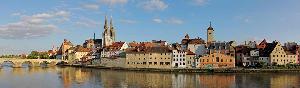A panorama of the city of regensburg from the river including a brick brdieg, a cathedral, old style houses and a clock tower.